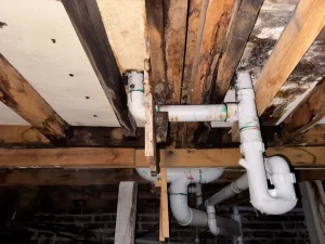 Close-up of water-damaged timber beams and white PVC plumbing pipes under a house, showing signs of leaks and rot.