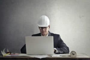 A man in business suit wearing a hard hat while working in front of a laptop