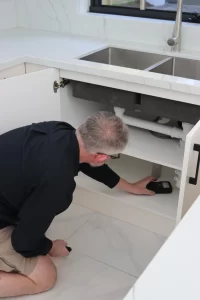 Pest control specialist inspecting under kitchen sink cabinets with flashlight to detect signs of pests.