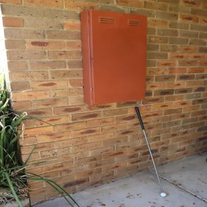 Orange metal utility or electrical box mounted on an exterior brick wall with a golf club leaning against the wall below it.