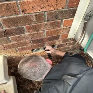 Building inspector using a flashlight to check subfloor ventilation vent in a brick wall.