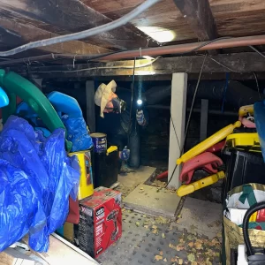 Building inspector wearing a respirator and using a flashlight while inspecting a cluttered subfloor crawl space.